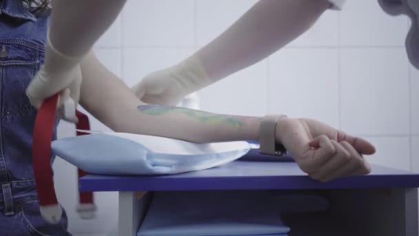 Detailní záběr lékaře připravujícího tetovanou pacientku na krevní testy. Nerozpoznatelná bělošská ošetřovatelka, která před vpichem obvazuje mladé ženy rukou. Lékařské vyšetření, Covid-19 pandemie. — Stock video