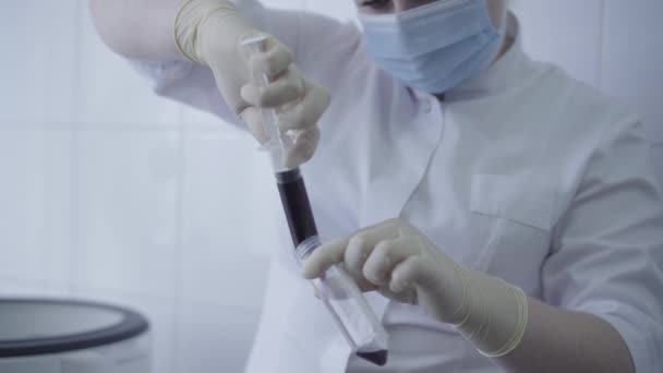 Серьёзный лаборант проливает кровь из шприца во фляжку для обработки в центрифуге. Белая женщина в маске изучает образцы пандемии Ковида-19. Медицина, лаборатории . — стоковое видео