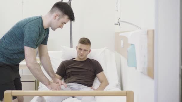 Young medical trainee examining ill knee joint of patient lying in hospital ward. Side view portrait of Caucasian brunette man consulting sportsman having trauma or injury. — Stock Video