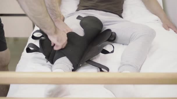 Mãos masculinas aplicando tala na perna do homem caucasiano ferido. Voluntário irreconhecível cuidando de paciente doente do sexo masculino na enfermaria. Reabilitação, tratamento, voluntariado . — Vídeo de Stock