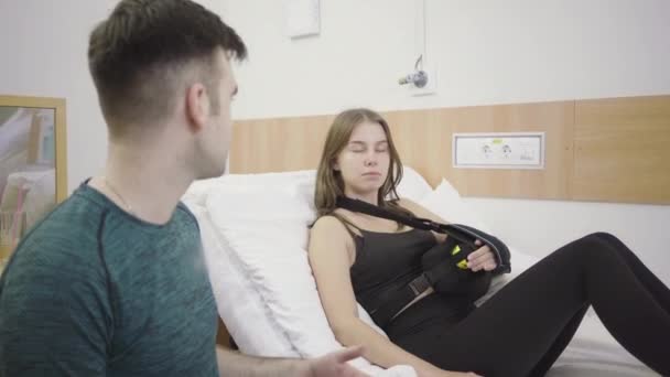 Portrét smutné unavené ženy se zlomenou rukou ležící na lůžku v nemocnici a poslouchající muže, jak mluví. Portrét mladé krásné bělošské dívky podstupující rehabilitaci po nehodě. Zdravotní péče — Stock video