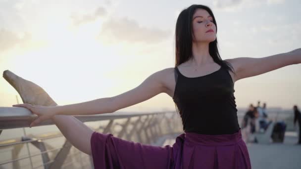 Prostřední záběr baletky, jak stojí rozpolcená a ohýbá se k noze. Portrét půvabné mladé brunetky běloška provádějící klasický baletní pohyb na mostě při východu slunce. — Stock video