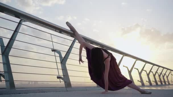 Bailarina profissional de pé em divisão na ponte com o sol brilhando no fundo. Retrato de tiro largo de dançarina de balé elegante caucasiana se movendo ao nascer do sol ao ar livre . — Vídeo de Stock