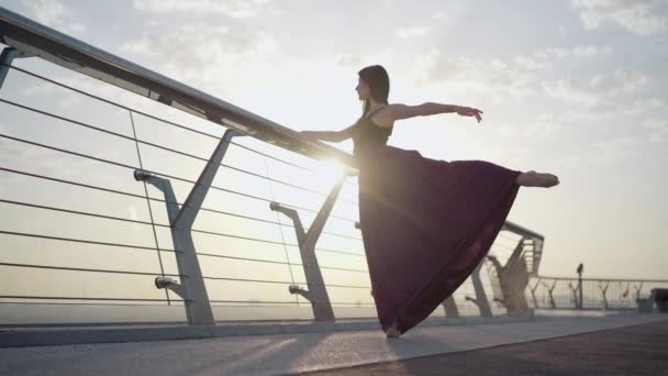 在阳光下,芭蕾舞演员在桥上跳舞的镜头很宽.自信优雅的高加索女人抬起腿，手拿着它。专业芭蕾舞演员在日出时在桥上练习.优雅的举止. — 图库视频影像