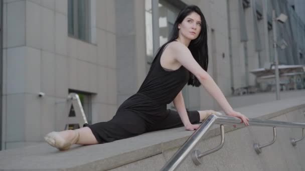 Вид сбоку портрет очаровательной молодой уверенной женщины, наклоняющейся вперед, сидящей в расщеплении. Великолепная кавказская балерина, выступающая на фоне современного городского здания . — стоковое видео