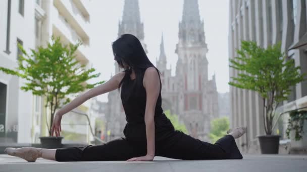 Jeune ballerine caucasienne mince assise en fente sur la rue urbaine et se penchant en arrière tenant le pied avec la main. Vue latérale d'une danseuse de ballet brune professionnelle dans une ville européenne avec des bâtiments anciens. — Video