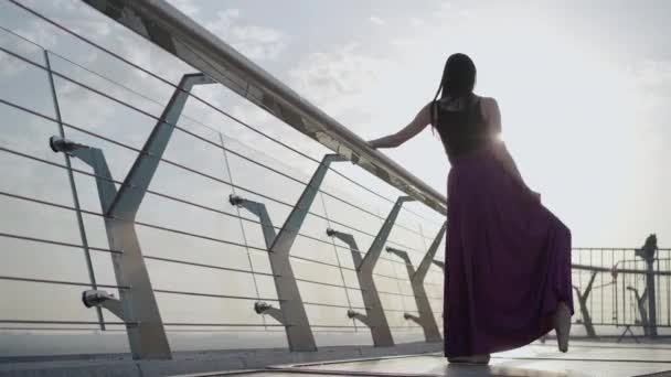 Vista posterior retrato de bailarina caucásica levantando el pie en los rayos del sol de la mañana. Amplio tiro de delgada mujer caucásica confiada de pie en split en puente al amanecer de verano . — Vídeos de Stock