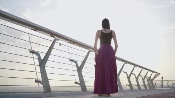 Силует Кавказької балерини рухає руками вгору і вниз сонячними променями. Широкий вид професійної танцівниці балету з літнім сходом сонця на мосту.. — стокове відео