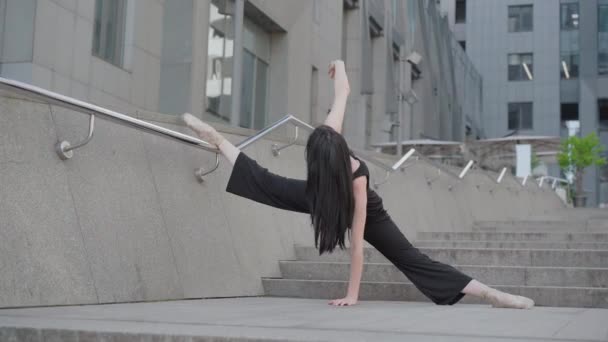 Brunette ballerine caucasienne debout divisé à l'arrière-plan d'un bâtiment moderne en ville et levant la main. Vue d'ensemble d'un danseur de ballet classique professionnel pratiquant à l'extérieur. Chorégraphie. — Video