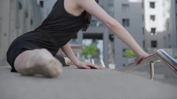 Slim nádherný balerína ohýbání na schodech zábradlí na městské městské ulici. Boční pohled na krásné bělošky sedí v split s kancelářskou budovou v pozadí. — Stock video
