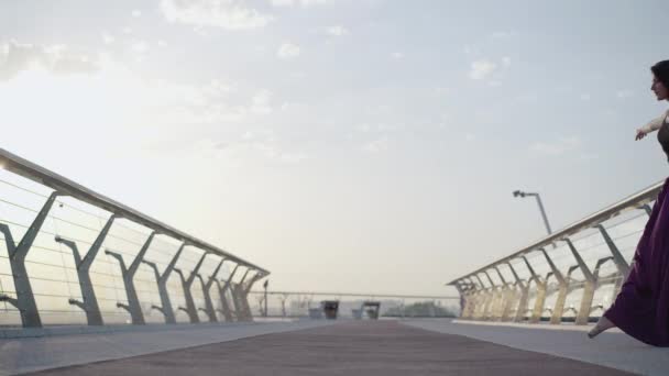 Bailarina caucásica saltando al amanecer y de pie en posición de ballet sobre puente. Vista lateral amplia toma de morena delgada mujer elegante bailando al aire libre en la mañana. Estilo de vida, encanto, elegancia . — Vídeo de stock