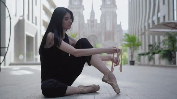 Seriöst elegant ballerina binda tåspetsskor sitter på asfalt med antika byggnader i bakgrunden. Sidovy porträtt av charmiga kaukasiska brunett kvinna i europeiska staden. — Stockvideo