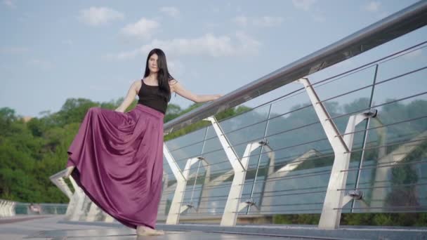 Breed schot van prachtige ballerina holding brug leuning en het verhogen van been omhoog. Portret van een zelfverzekerde blanke balletdanser die buiten oefent op zonnige zomerdag. Lifestyle, elegantie. — Stockvideo