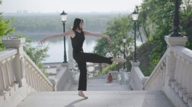 Slim balerin güneşli bir günde şehir merdivenlerinde bale yapıyor. Açık havada dans eden güzel, kendine güvenen beyaz kadın portresi. Dansçı hobisinin geniş açılı görüntüsü..