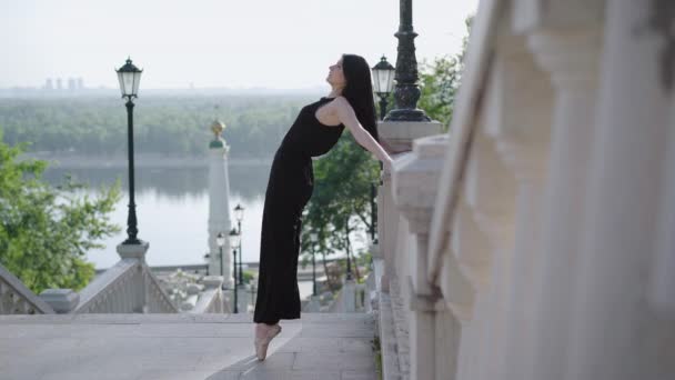 Vista lateral de la encantadora bailarina elegante inclinándose hacia atrás de pie en un dedo del pie. Amplio plano de caucásico morena mujer bailando en las escaleras urbanas con coches a caballo en el fondo . — Vídeo de stock