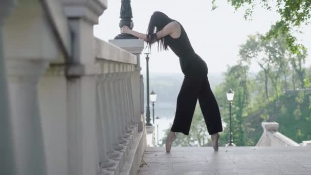 Amplio plano de bailarina en traje negro que se extiende en las escaleras urbanas. Vista lateral de la delgada morena caucásica mujer de pie en puntillas. Elegante bailarina de ballet que practica al aire libre en un día soleado . — Vídeos de Stock