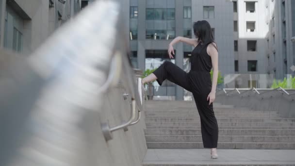 Zijaanzicht van zelfverzekerde ballerina in zwart kostuum staan op de trap op de achtergrond van de moderne stedelijke gebouw en verplaatsen. Breed shot portret van professionele mooie blanke vrouw. — Stockvideo