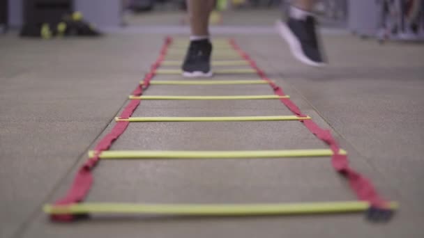 スニーカーを着た男性の足は、ジムの床に横たわってはしごを踏んでいます。認識できない白人スポーツマンのトレーニング室内のクローズアップ足。運動、スポーツ、健康的なライフスタイルの概念. — ストック動画