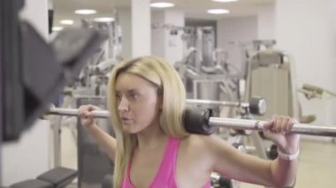 Kamera, omuzlarında ağırlıklarla çömelmiş kendine güvenen bir kadının hareketini takip ediyor. Spor salonunda envanterle antrenman yapan güzel sarışın beyaz kadın portresi. Sportif yaşam tarzı.