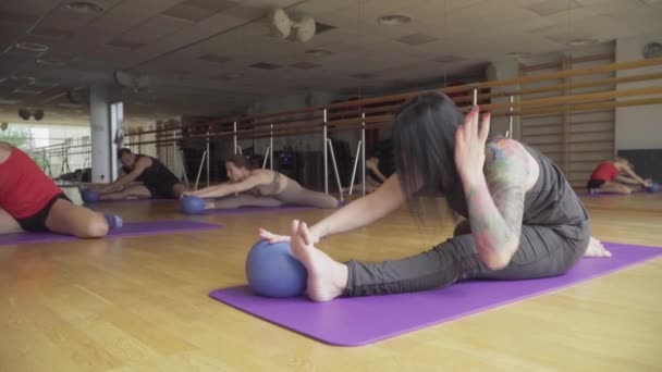Breed shot van zelfverzekerde atletische mensen die yoga beoefenen in de sportschool. Sportmannen en -vrouwen die op een yogamat zitten en naar voren buigen om met de bal in de hand te spelen. Workout, sport, levensstijl. — Stockvideo