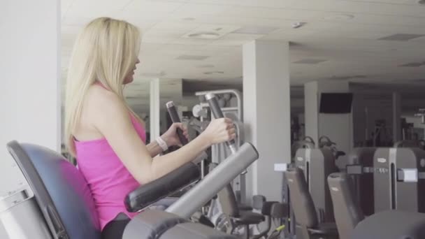 Vista lateral da mulher loira confiante levantando pernas no ginásio. Jovem bela esportista caucasiano músculos de treinamento no inventário desportivo. Slim menina atraente exercitar-se em equipamentos no clube de esportes . — Vídeo de Stock