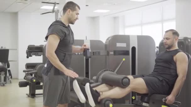 Jovem desportista focado levantando pesos de perna com a ajuda de treinador pessoal. Treinamento de instrutor de fitness Homem caucasiano no ginásio. Aptidão, masculinidade, desporto, estilo de vida . — Vídeo de Stock