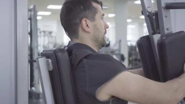 Close-up zijaanzicht van jonge geconcentreerde man die traint op fitnesstoestellen. Portret van een sterke blanke sportman die trainingsapparatuur gebruikt voor handspieren. Sport, mannelijkheid, kracht. — Stockvideo