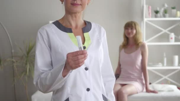 Médico caucásico irreconocible mostrando agujas para la acupuntura en la cámara como mujer rubia borrosa sentada en el sofá en el fondo y sonriendo. Retrato de acupunturista profesional de confianza . — Vídeo de stock