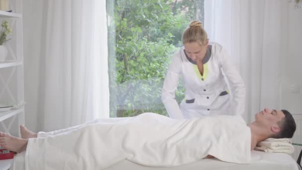 Médico profesional serio masajeando cuerpo de joven morena hombre cubierto con toalla blanca. Vista lateral panorámica del paciente varón caucásico acostado en el sofá en la clínica de acupuntura. Medicina, masaje . — Vídeos de Stock