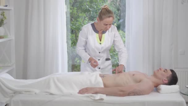 O lovitură largă de medic profesionist care introduce ace în burta tânărului om slab. Serios încrezător caucazian femeie acupuncturist tratarea bolnav pacient de sex masculin. Medicină alternativă, acupunctură . — Videoclip de stoc