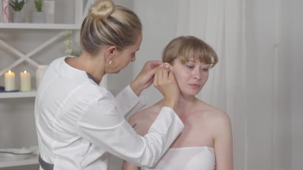 Middle shot of professional serious acupuncturist inserting needles into ear of concentrated Asian woman. Doctor treating young slim girl. Traditional chinese acupuncture. — Stock Video