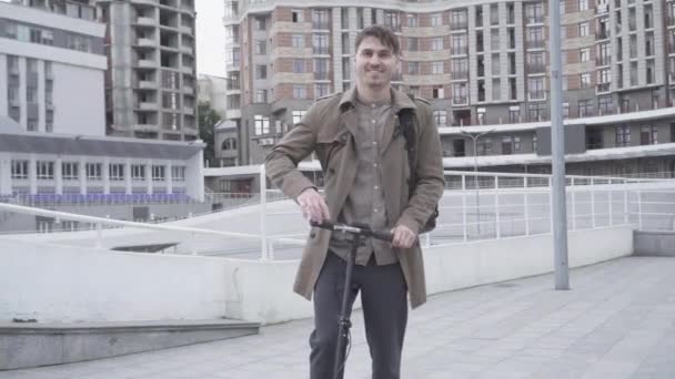 Portret van een vrolijke volwassen blanke man die op straat staat met een scooter en glimlacht naar de camera. Jonge brunette die plezier heeft buiten. Individualiteit, levensstijl, geluk. — Stockvideo
