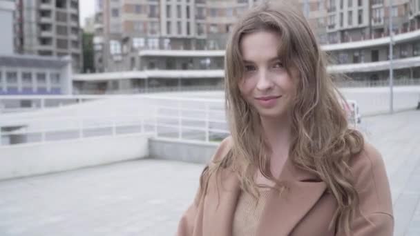 Ritratto ravvicinato di affascinante ragazza bruna dagli occhi castani che guarda la fotocamera e sorride. Bella giovane donna caucasica in posa all'aperto sulla strada della città il giorno ventoso. Stile di vita concetto. — Video Stock