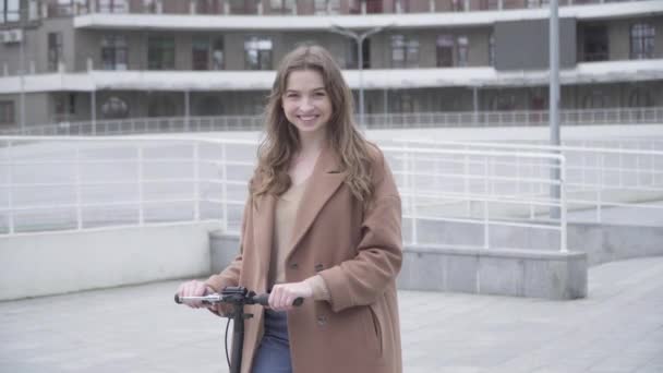 Portret van lachend mooi meisje poseren met scooter buiten op bewolkte dag. Midden schot van positieve jonge blanke vrouw genieten van hobby op stedelijke straat. Vreugde, levensstijl, vrije tijd. — Stockvideo