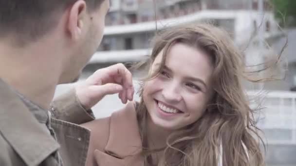 Retrato de rir menina morena feliz namoro ao ar livre com namorado. Amoroso jovem caucasiano ajustando namoradas cabelo soprado com vento. Casal alegre desfrutando de seu amor . — Vídeo de Stock