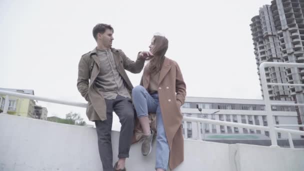 Amplio plano de alegre pareja joven sentada al aire libre en la ciudad y charlando. Retrato de un hombre y una mujer caucásicos felices hablando y riendo en un día nublado. Romance, amor, citas, felicidad . — Vídeos de Stock