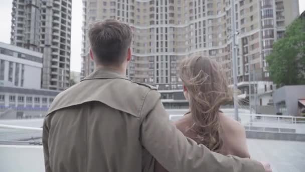 Брюнетка молодая женщина кладет голову на плечо уверенного мужчины, обнимающего ее на улице. Задний вид молодой кавказской пары, стоящей на улице в ветреный день и смотрящей на небоскреб . — стоковое видео