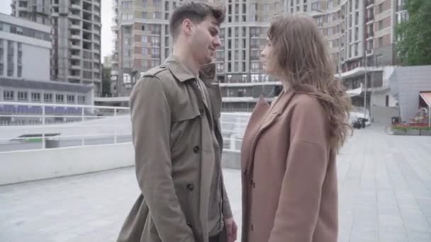 Kameran närmar sig en ung man som kysser en kvinna på en blåsig gata. Sidovy porträtt av unga kaukasiska par med datum utomhus. Begreppet första kyss, kärlek, romantik. — Stockvideo
