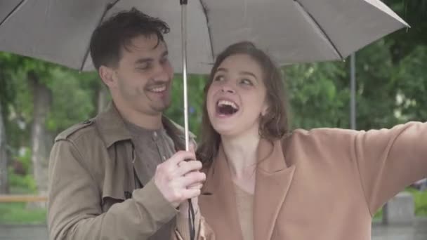 Retrato de cerca de una alegre pareja caucásica riéndose de pie bajo el paraguas en un día lluvioso. Feliz novio sonriente y novia disfrutando de citas bajo la lluvia al aire libre. Amor, felicidad, alegría . — Vídeos de Stock
