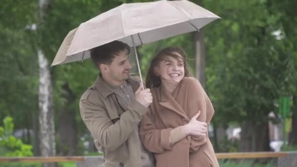 Retrato de homem bonito caucasiano positivo sorrindo e abraçando namorada amorosa no dia chuvoso ao ar livre. Meio tiro de feliz jovem casal caucasiano em pé na rua da cidade sob guarda-chuva e rindo — Vídeo de Stock