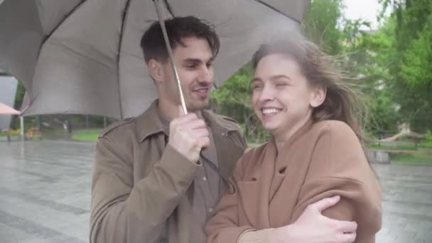 Pria muda yang tampan mencium wanita yang bahagia Menaruh kepalanya di bahunya. Potret pasangan bahagia Kaukasia yang berkencan di jalan kota pada hari hujan mendung. — Stok Video