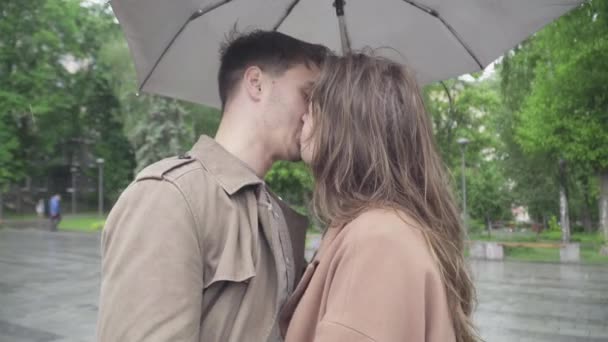 Vista lateral de cerca de la pareja amorosa besándose bajo el paraguas en el día lluvioso. Retrato de hombre y mujer caucásica romántica feliz citas en la calle de la ciudad bajo la lluvia. Alegría, ocio, amor, estilo de vida . — Vídeos de Stock