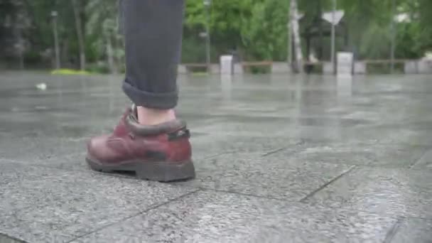 Cámara sigue los pasos de elegante joven hombre caucásico. Primer plano de los pies masculinos en botas caminando sobre el pavimento a lo largo de la calle urbana de la ciudad. Un hombre irreconocible paseando al aire libre en un día lluvioso . — Vídeo de stock