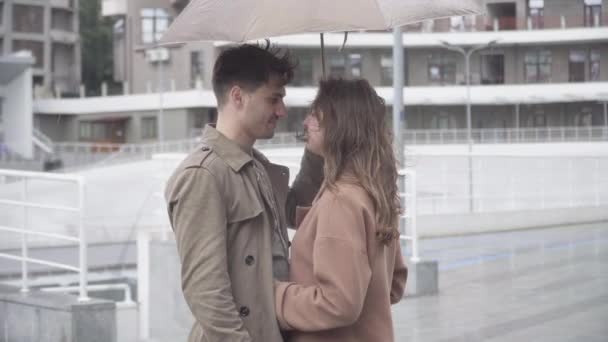 Молодий чоловік і жінка ховаються за парасолькою, щоб поцілуватися. Бічний вид середнього виду на люблячу кавказьку пару, що зустрічається на відкритому повітрі в місті в дощовий день. Любов, романтика, зв'язок, єдність. — стокове відео