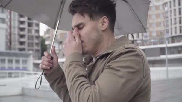 Närbild av unga sjuka kaukasiska mannen står utomhus med paraply, nysningar och hosta. Sidovy porträtt av stilig brunett kille väntar på möte på urban city street. Hälsovård, Covid-19. — Stockvideo