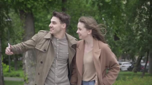 Positivt kaukasiskt par som tar selfie utomhus på regnig dag. Porträtt av glada unga kaukasiska man och kvinna att göra ansikten som tittar på kameran på smartphone. Dejting, fritid, nöje, livsstil. — Stockvideo