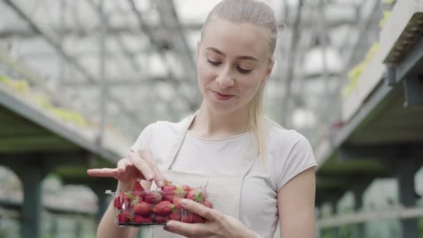 Cámara se acerca a la cara hermosa de la joven mujer caucásica mordiendo fresa dulce y sonriendo a la cámara. Retrato de chica blanca encantadora positiva comiendo baya orgánica en invernadero . — Vídeos de Stock