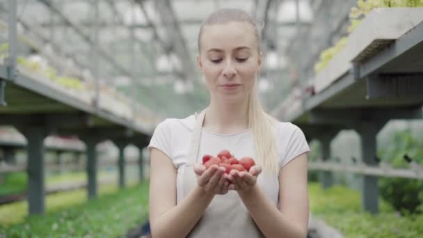Charmante junge Kaukasierin mit Erdbeere in der Hand und Blick in die Kamera. Porträt einer schönen Biologin, die im Gewächshaus posiert. Lifestyle, Schönheit. — Stockvideo