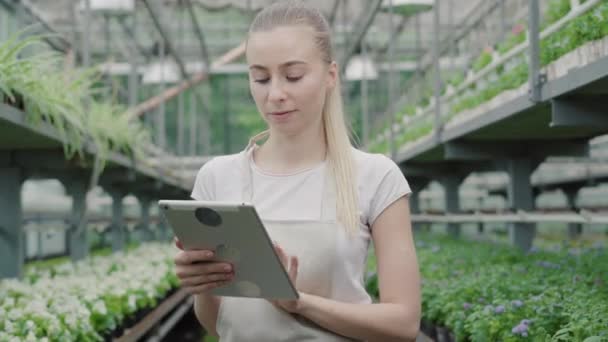 Kamera nähert sich charmanter blonder Frau mit Tablette im Gewächshaus. Schöne junge kaukasische Agronomin oder Biologin, die im Treibhaus arbeitet. Hübsches Mädchen blickt in die Kamera und lächelt. — Stockvideo