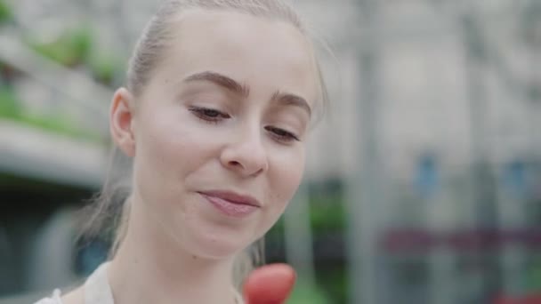 Närbild av blond vacker kvinna med blå grå ögon biter jordgubbe och ler mot kameran. Porträtt av charmig ung flicka som äter bär i växthus. Ekologisk mat, livsstil. — Stockvideo